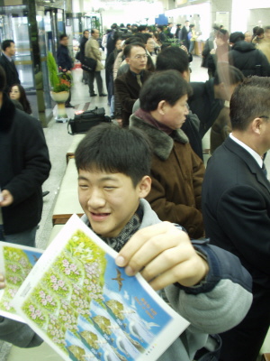 한 소년이 독도우표를 들고 웃고 있다. 뒤 편으로 독도우표를 사기위해 길게 늘어선 줄이 보인다 